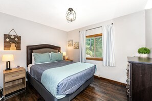A bedroom with a bed, bedside tables, a lamp, a clock, a window with curtains, and a wall-mounted light fixture.