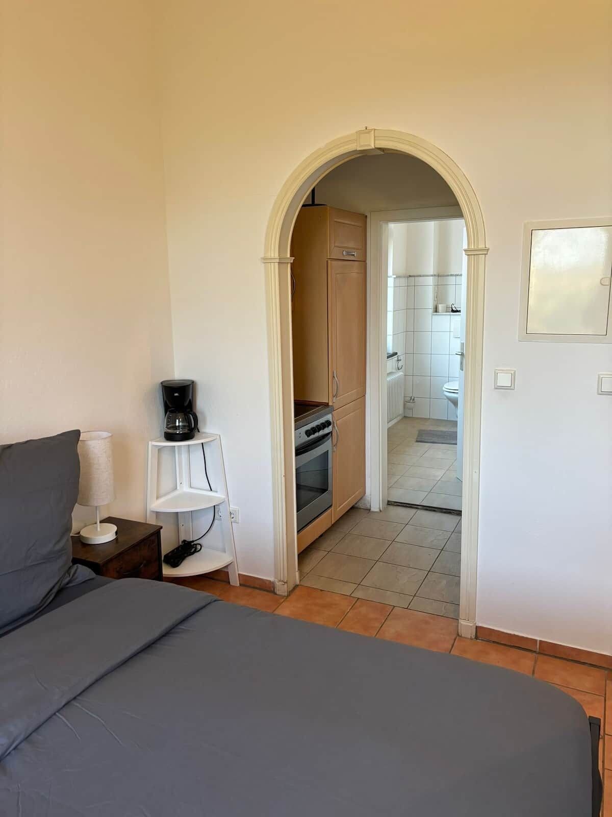 Een slaapkamer met een bed, een koffiezetapparaat en een plank. Er is een boog die leidt naar een keuken met een fornuis en een spoelbak.