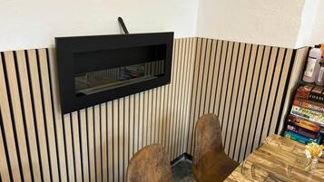 Een hoek van een kamer met een raam met donker frame, een wand van houten latten, twee houten stoelen en een klein tafeltje met boeken.
