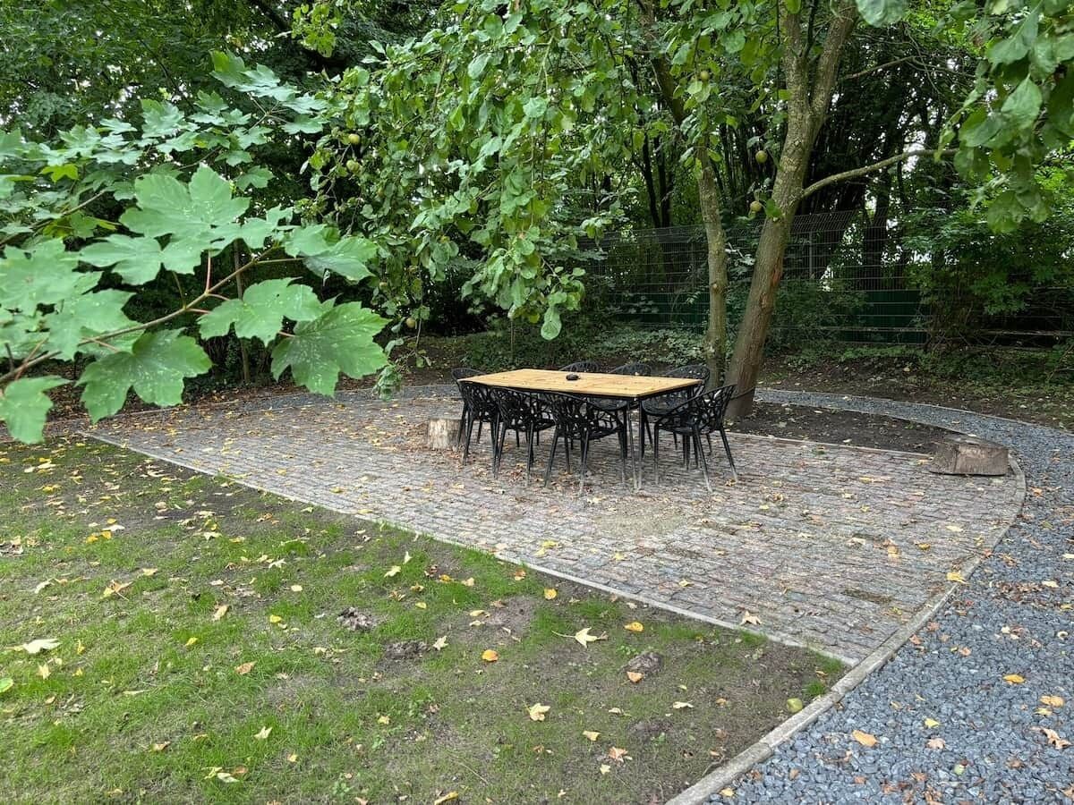 An outdoor seating area with a table and chairs, surrounded by trees and a brick pathway.
