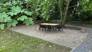 An outdoor seating area with a table and chairs, surrounded by trees and a brick pathway.