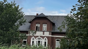 A two-story brick house with a gabled roof, surrounded by overgrown vegetation.