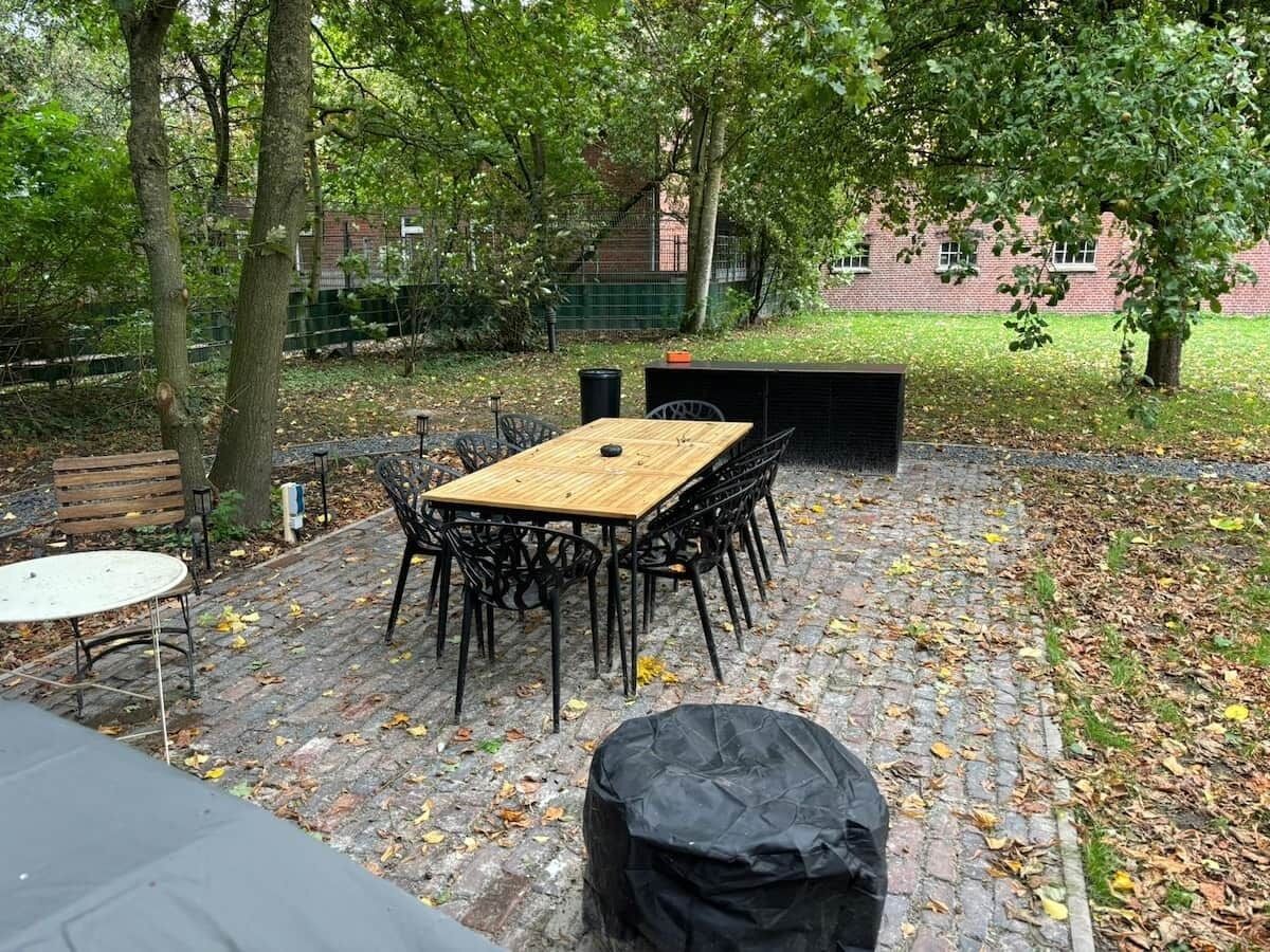 Une terrasse extérieure avec une table et des chaises en bois, entourée d’arbres et d’un mur de briques.