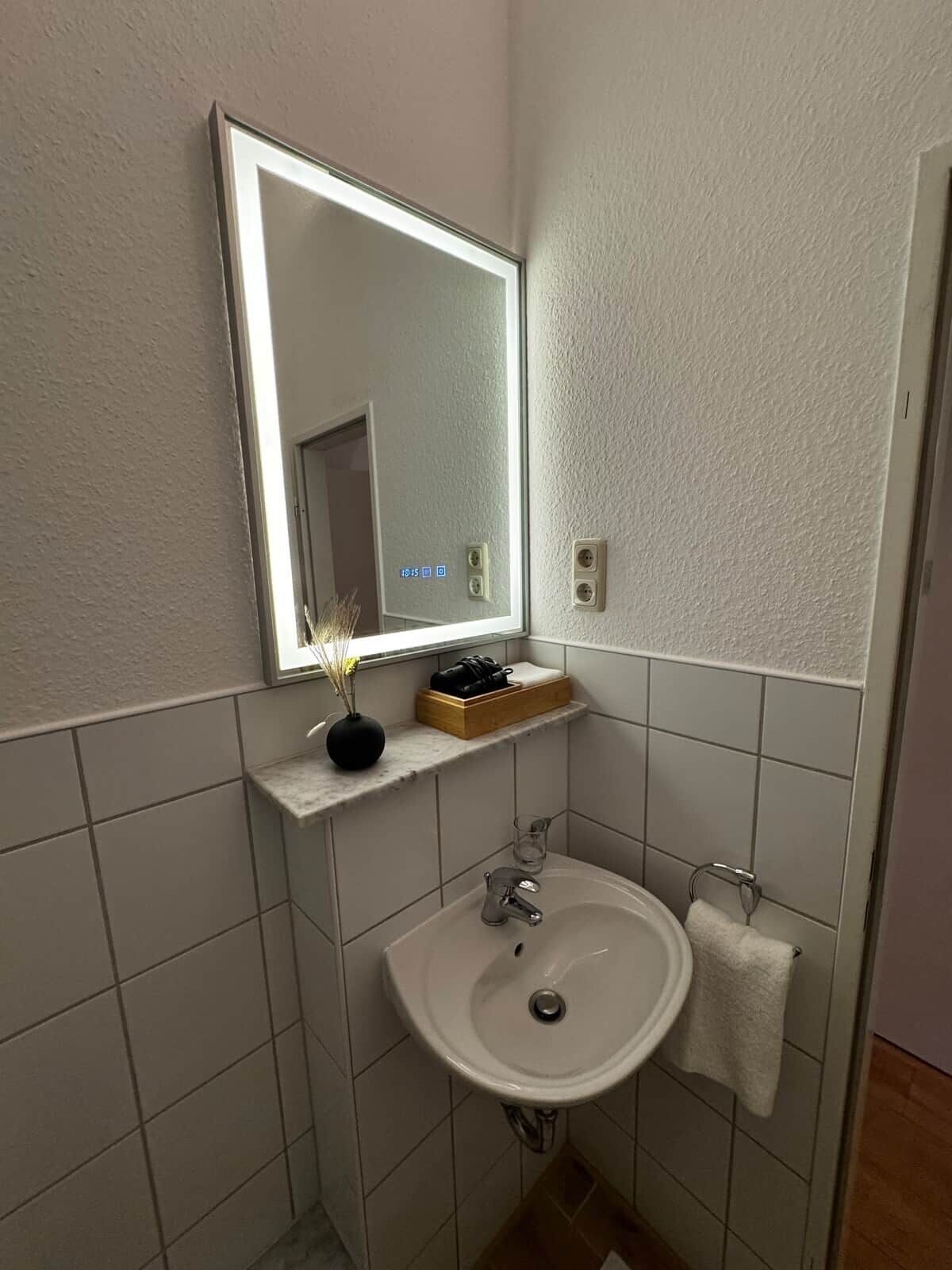 Un baño con un lavamanos blanco, un espejo, un perchero para toallas y un estante con un jarrón y una caja.