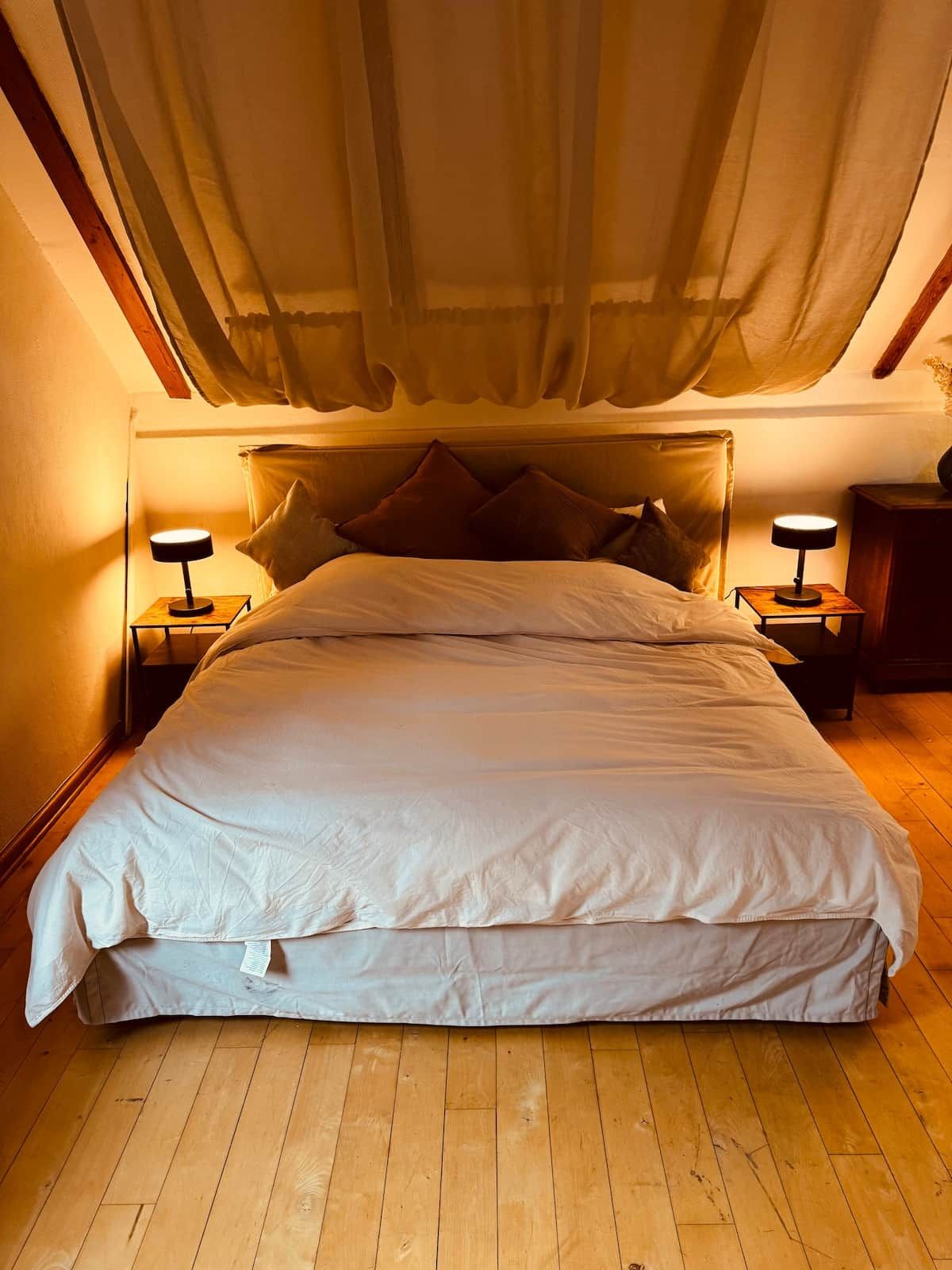 A bed with white bedding, two bedside tables with lamps, a wooden floor, and a sheer curtain above.