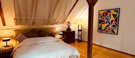 Un dormitorio con una cama grande, vigas de madera, una mesita de noche, una lámpara y un cuadro con motivos geométricos en la pared.