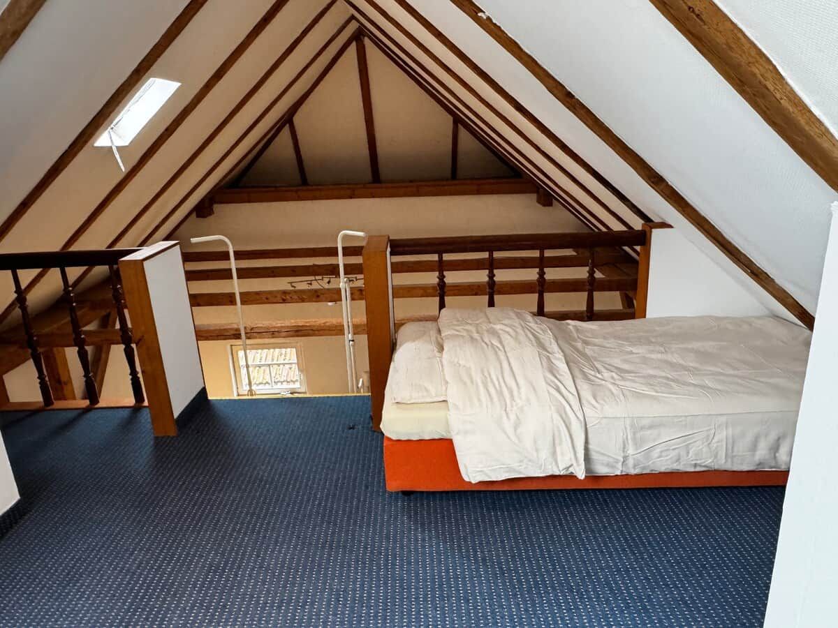 Ein Schlafzimmer mit schrägen Wänden, einem Bett, einer Leiter und einem Fenster.