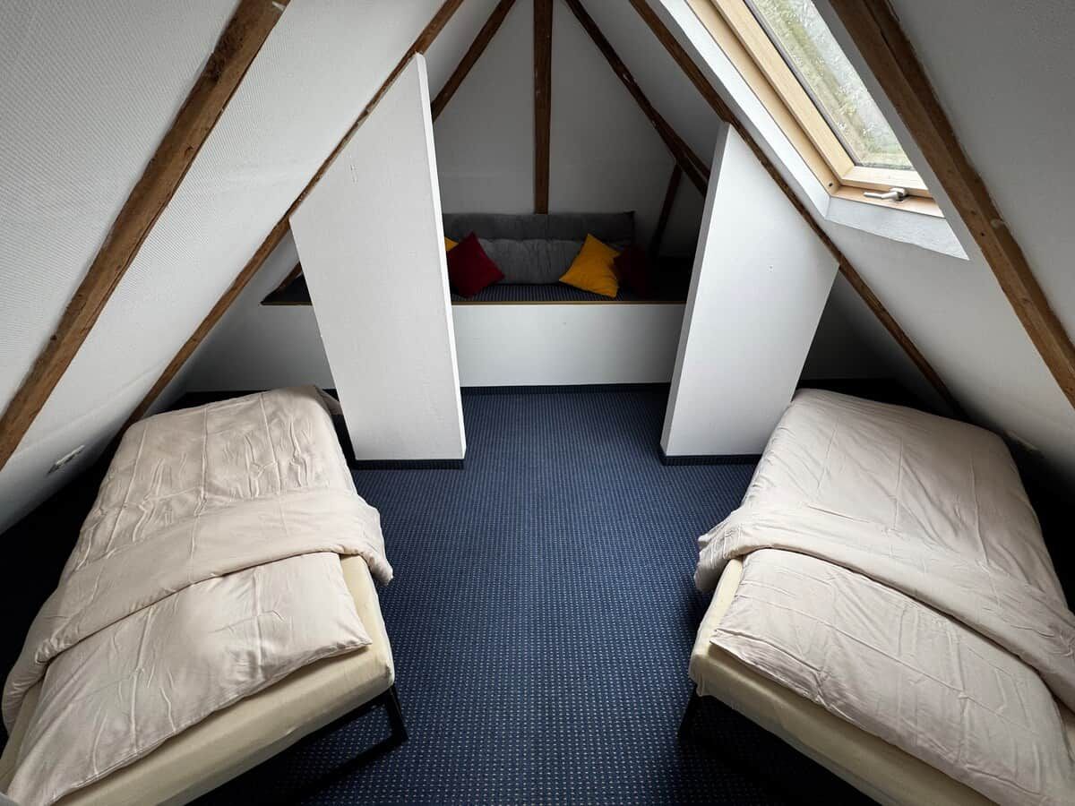 Ein kleines Schlafzimmer im Dachgeschoss mit zwei Betten, einem Fenster und einer eingebauten Sitzbank mit Kissen.