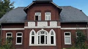 A two-story brick house with a gabled roof, a balcony with ornate railings, and three arched windows on the second floor.