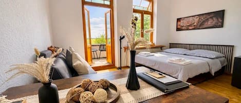 A bedroom with a bed, a table with decorative items, and a view through the open window.