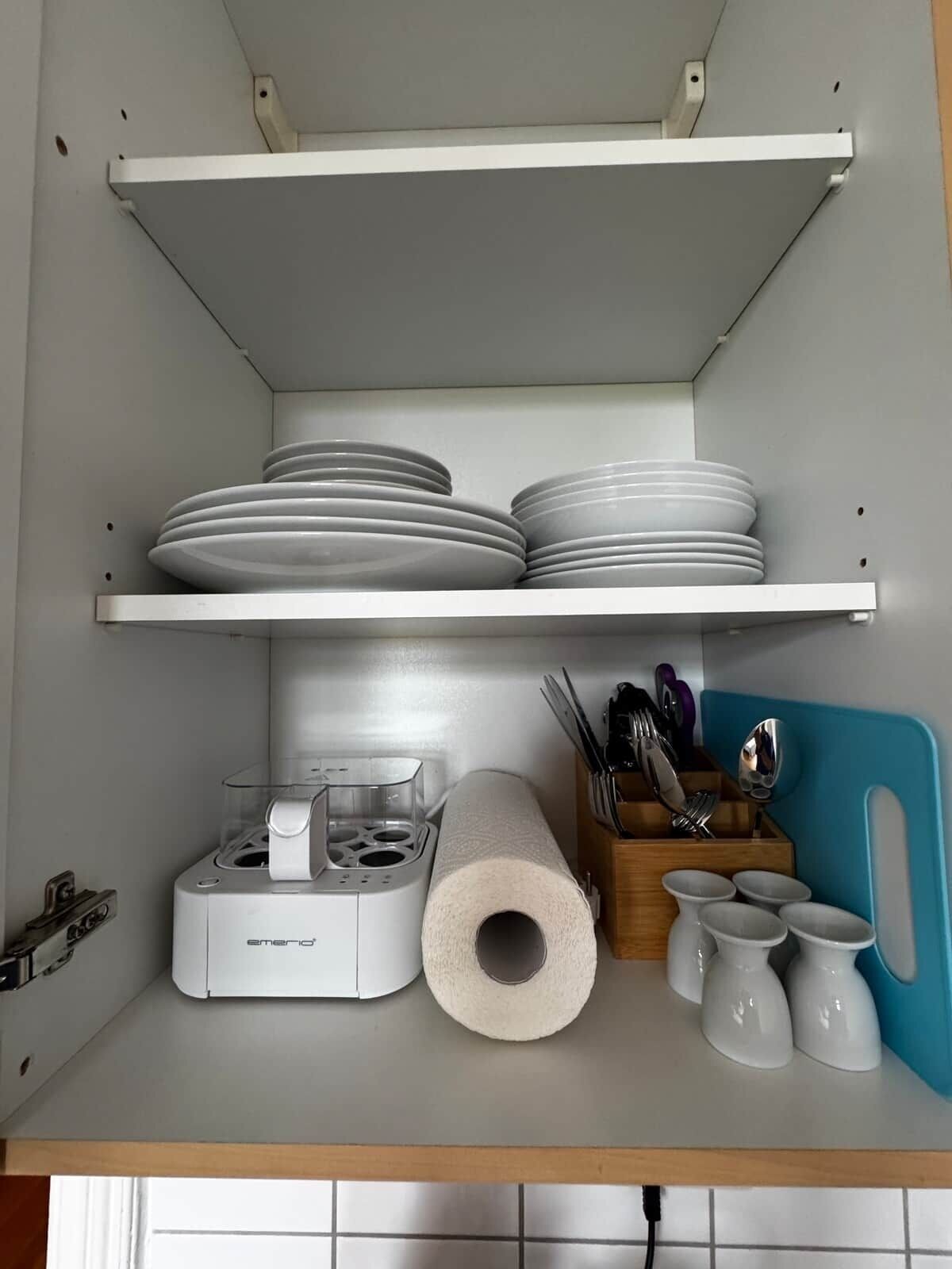 Ett kök med vita hyllor, en kaffebryggare, en rulle hushållspapper och köksredskap.