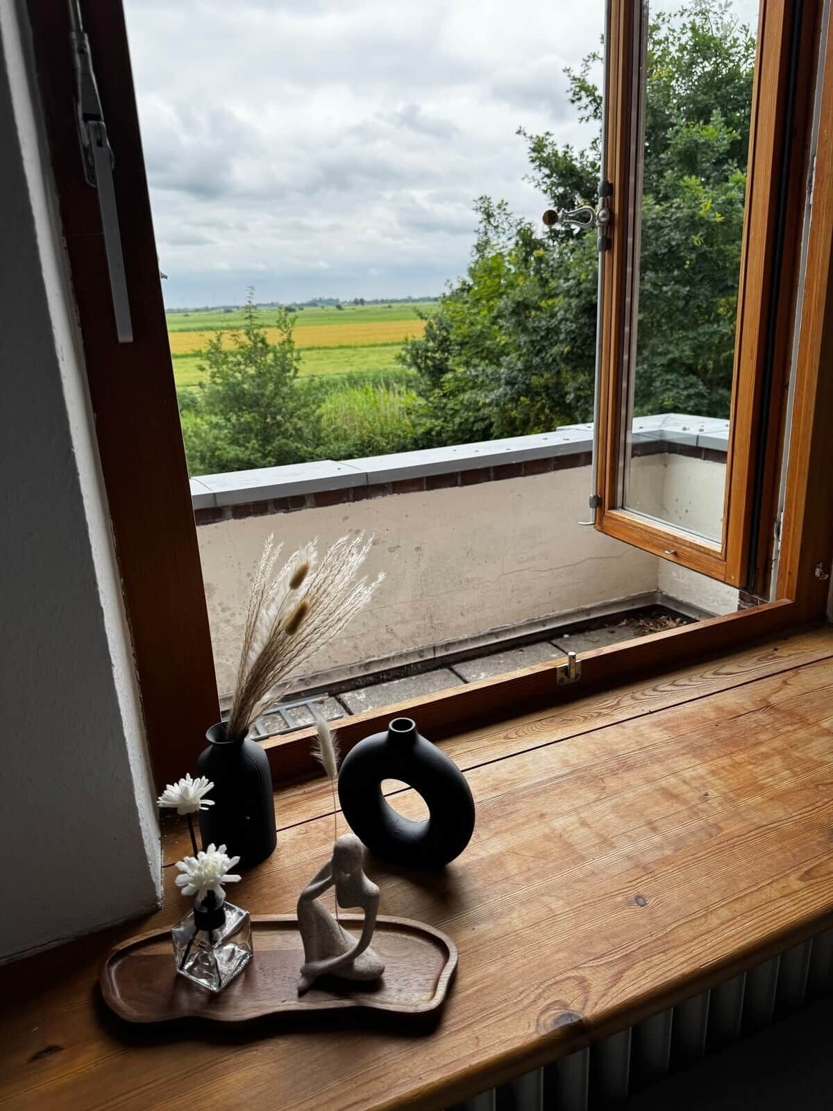 A window with a view of a green field and trees, a wooden bench, and two vases with dried plants.