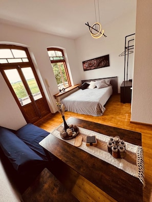Ein Schlafzimmer mit einem Bett, einer Bank, einem Tisch mit Dekoration, einem Gemälde und einem Fenster mit Aussicht.