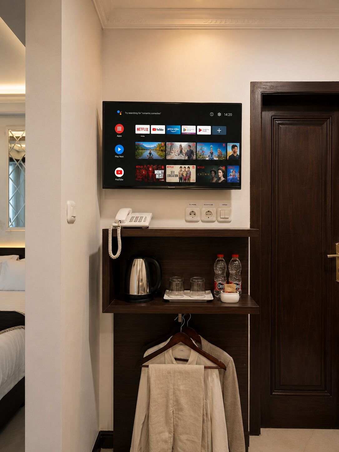 A hotel room with a mounted TV displaying various streaming apps, a telephone, a coffee maker, and a shelf with a kettle, glasses, and a bottle.