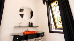 Un baño con un espejo redondo, tocador negro, plato para jabón naranja y una ventana de vidrio emplomado.