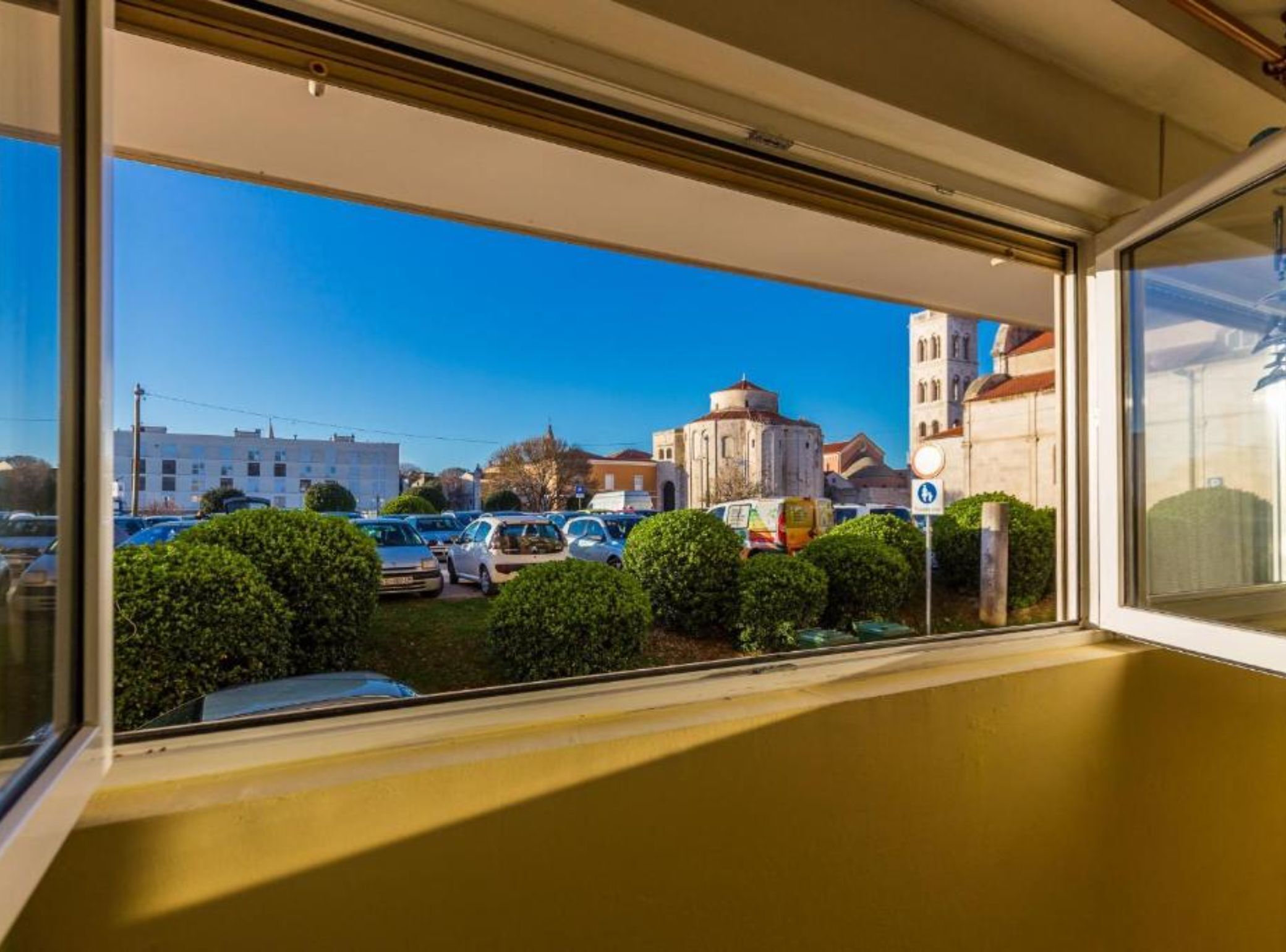Vista desde una ventana: cielo despejado, estacionamiento y edificios, incluyendo una iglesia con cúpula.