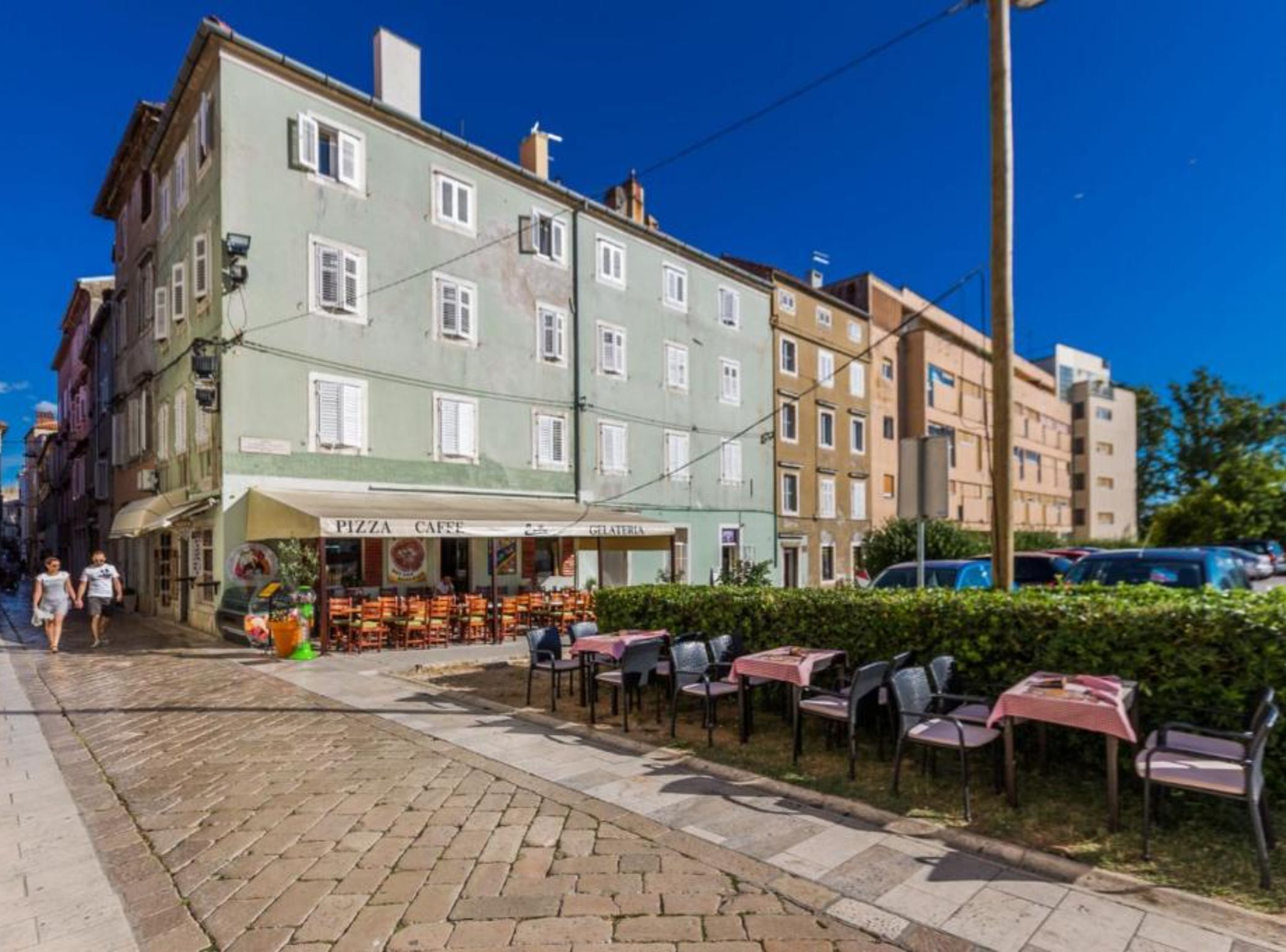 Ένας δρόμος με ένα καφέ που ονομάζεται «PIZZA CAFÉ» και διαθέτει τραπέζια για εξωτερική χρήση.