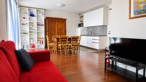 A modern living room with a red sofa, wooden dining table, and chairs, a kitchen area with white cabinets, and a flat-screen TV. - Elegant apartment with a garage, two bedrooms, two bathrooms, and two terraces (Alba)