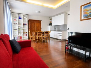 Ein modernes Wohnzimmer mit einem roten Sofa, einem Holzespeisetisch und Stühlen, einer Küchenzeile mit weißen Schränken und einem Flachbildfernseher.