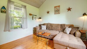 A living room with a sofa, a small table, a window with curtains, a wall clock, and a poster. - Cast & Relax! Wolf Creek Fishing Cabin w/ Dock (Grove)