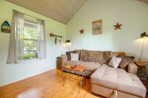 Ein Wohnzimmer mit einer Couch, einem kleinen Tisch, einem Fenster mit Vorhängen, einer Wanduhr und einem Poster.