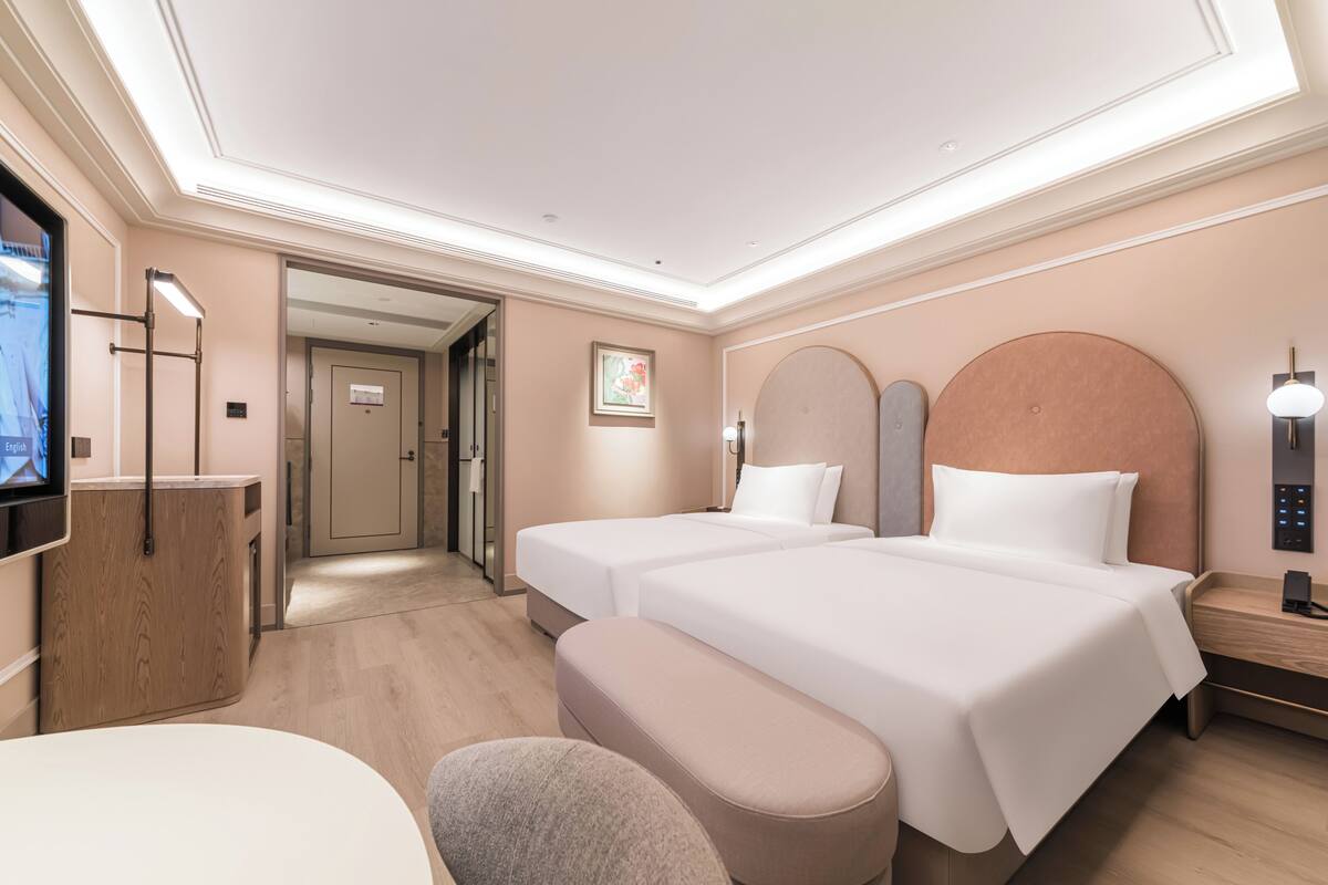 A hotel room with two beds, a desk, a chair, a TV, and a bathroom visible in the background.