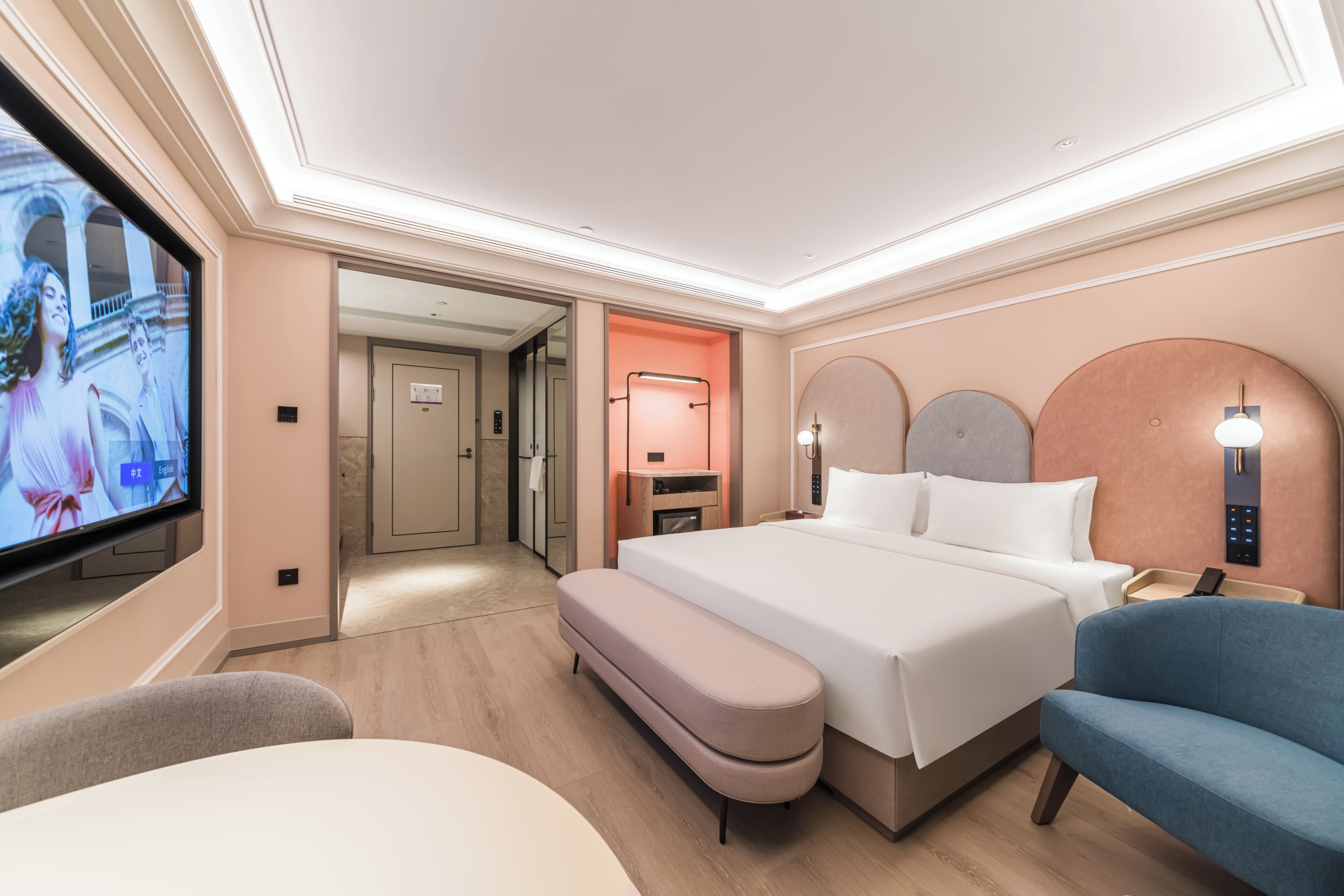 Habitación de hotel con cama, televisión, banca, silla y baño visible a través de una puerta abierta.