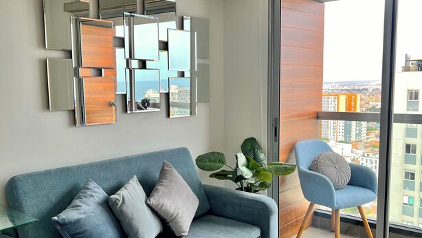 A modern living room with a blue sofa, a glass coffee table, and a city view through a sliding glass door.