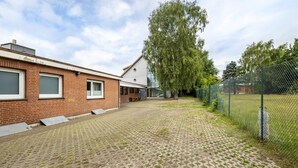 A courtyard with a brick building, a large tree, and a chain-link fence. - Apartment 'Zwei Betten Studio' with Wi-Fi (Lübeck)