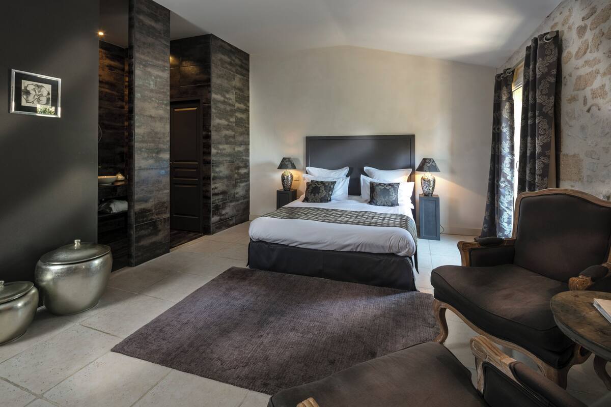 Un dormitorio con cama, lámparas de noche, una silla, un tapete y una mesita.