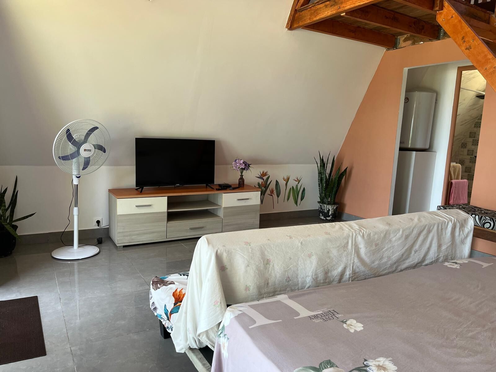 Ein Schlafzimmer mit einem Bett, einem Fernseher auf einem Holzständer, einem Standventilator und einer Topfpflanze.