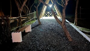 A nighttime view of a triangular building with a lighted entrance, surrounded by trees and gravel. - FAMILY BUNGALOW: E-FRAME HOUSE Facing the Ocean (Toamasina)