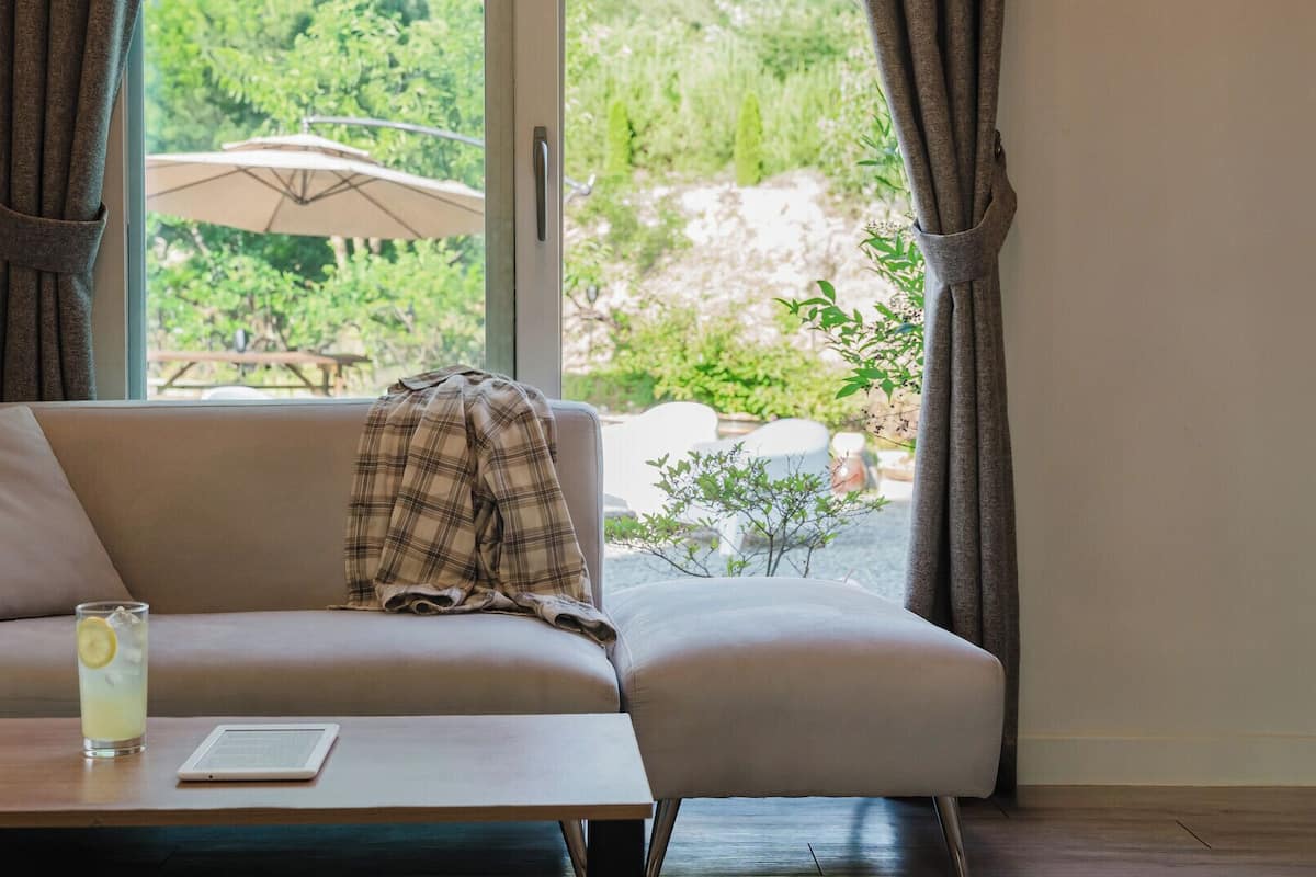 Una sala de estar moderna con un sofá, una mesa con una tableta y un vaso de agua, y vistas a un jardín a través de grandes ventanales.
