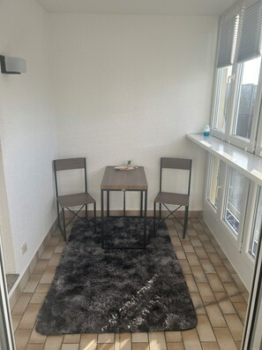 Ein Eckzimmer mit gefliestem Boden, einem schwarzen, flauschigen Teppich, einem kleinen Tisch mit zwei Stühlen und einem Fenster mit Jalousien.