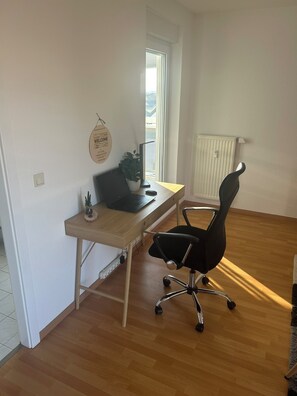 Ein Zimmer mit einem Holztisch, einem schwarzen Bürostuhl, einem Laptop, einer Topfpflanze und einer Heizung.