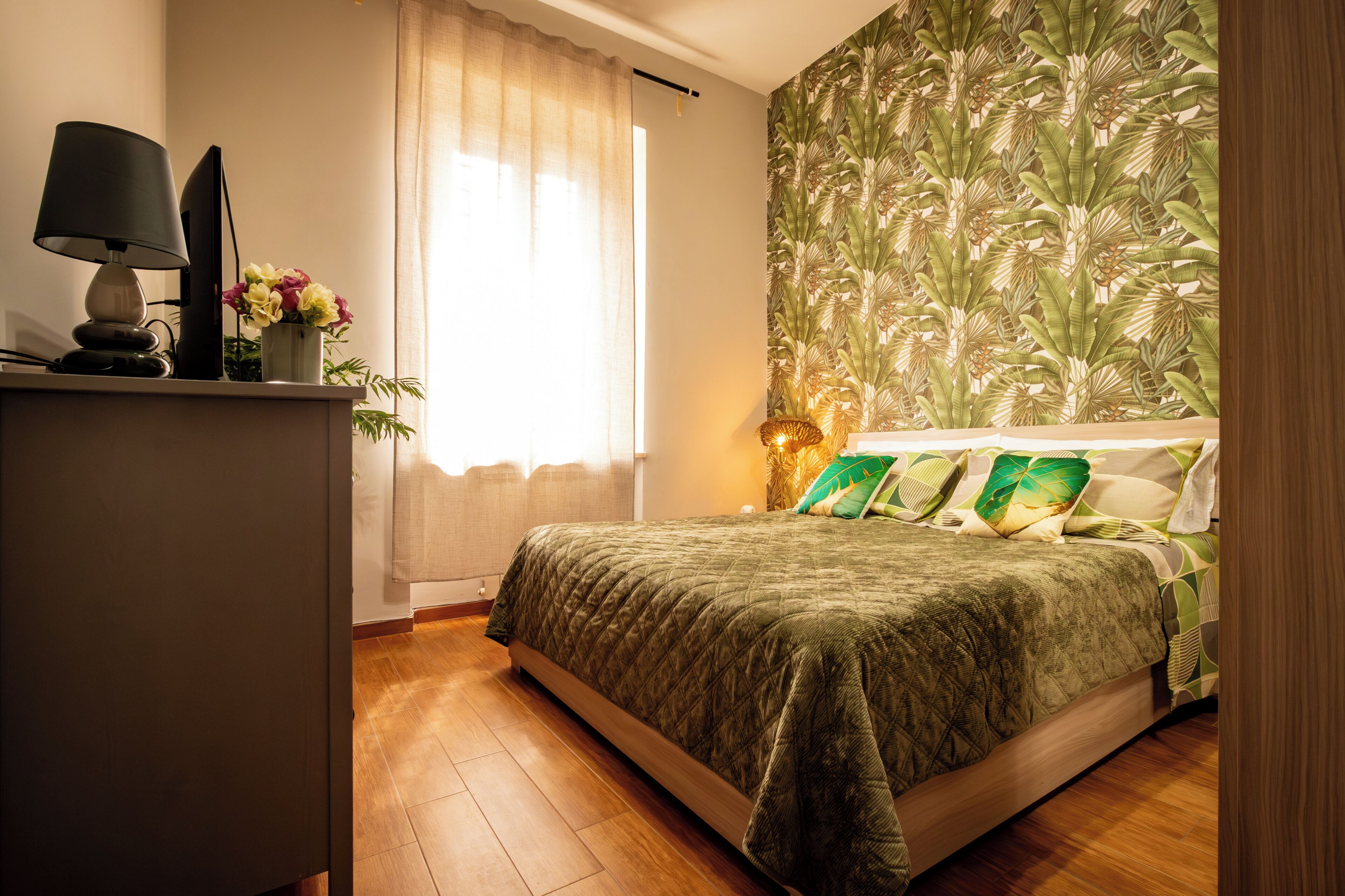 Ein Schlafzimmer mit einem Bett, einem Schreibtisch mit Lampe, einem Fenster mit Vorhängen und einer Wand mit tropischem Wandpapier.