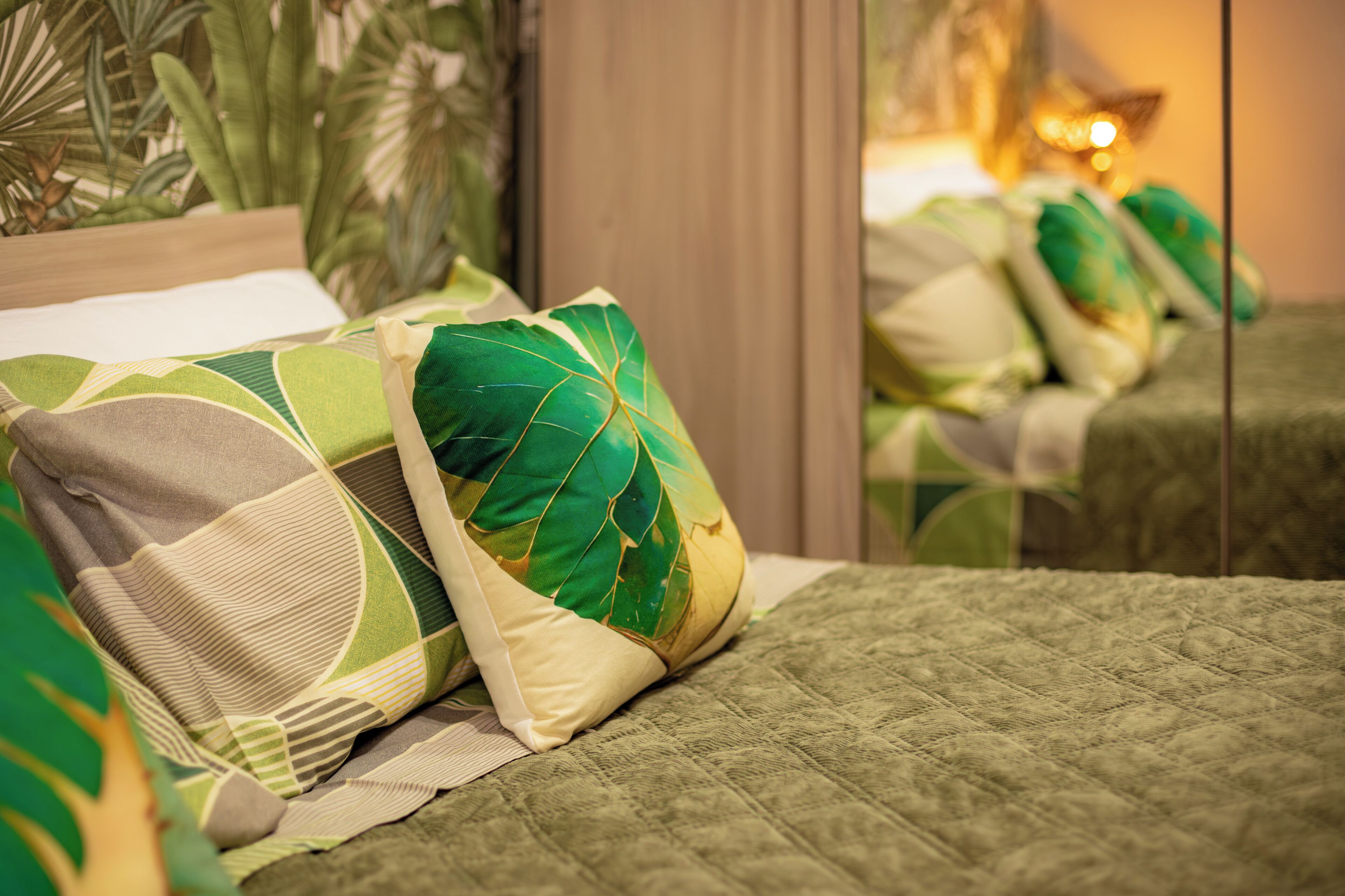 A bed with a green quilt, a pillow with a leaf pattern, and a mirror reflecting the room's interior.