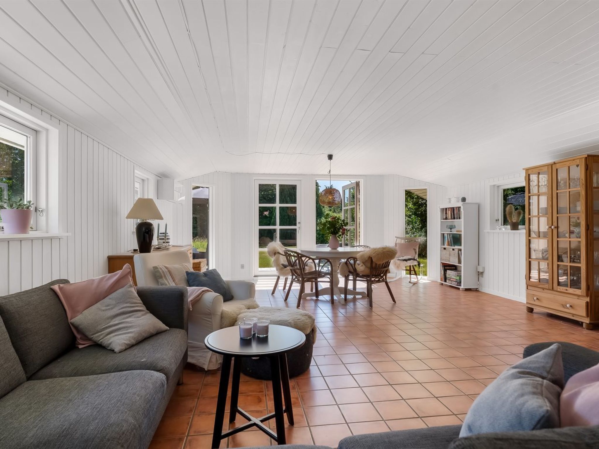 A spacious living area with a sofa, a round table, and a dining table with chairs. There is a cabinet with glass doors and a window with a view of greenery.