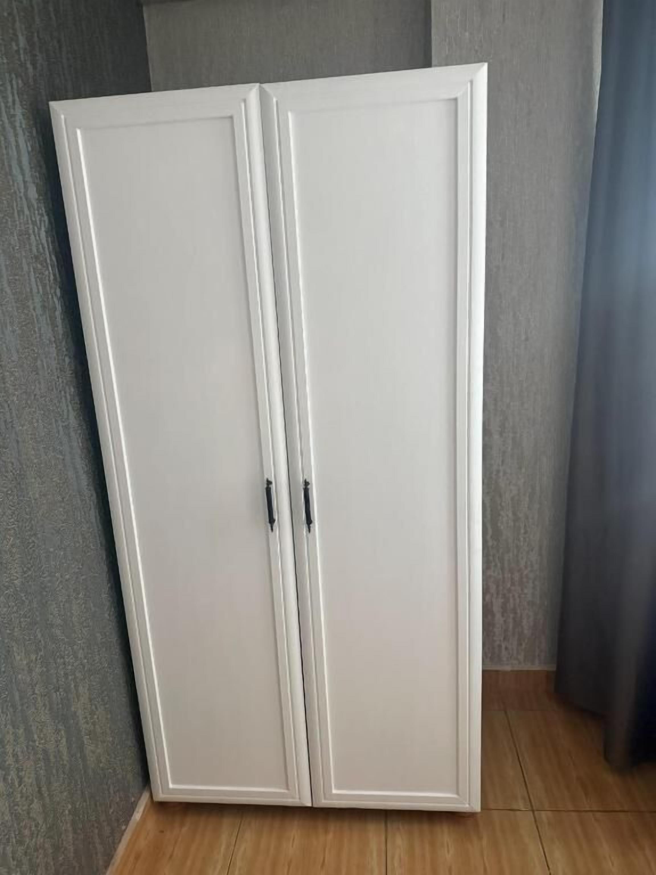 A white wardrobe with two doors and a handle, against a grey wall and wooden floor.