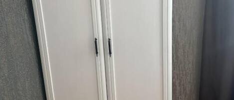 A white wardrobe with two doors and a handle, against a grey wall and wooden floor.