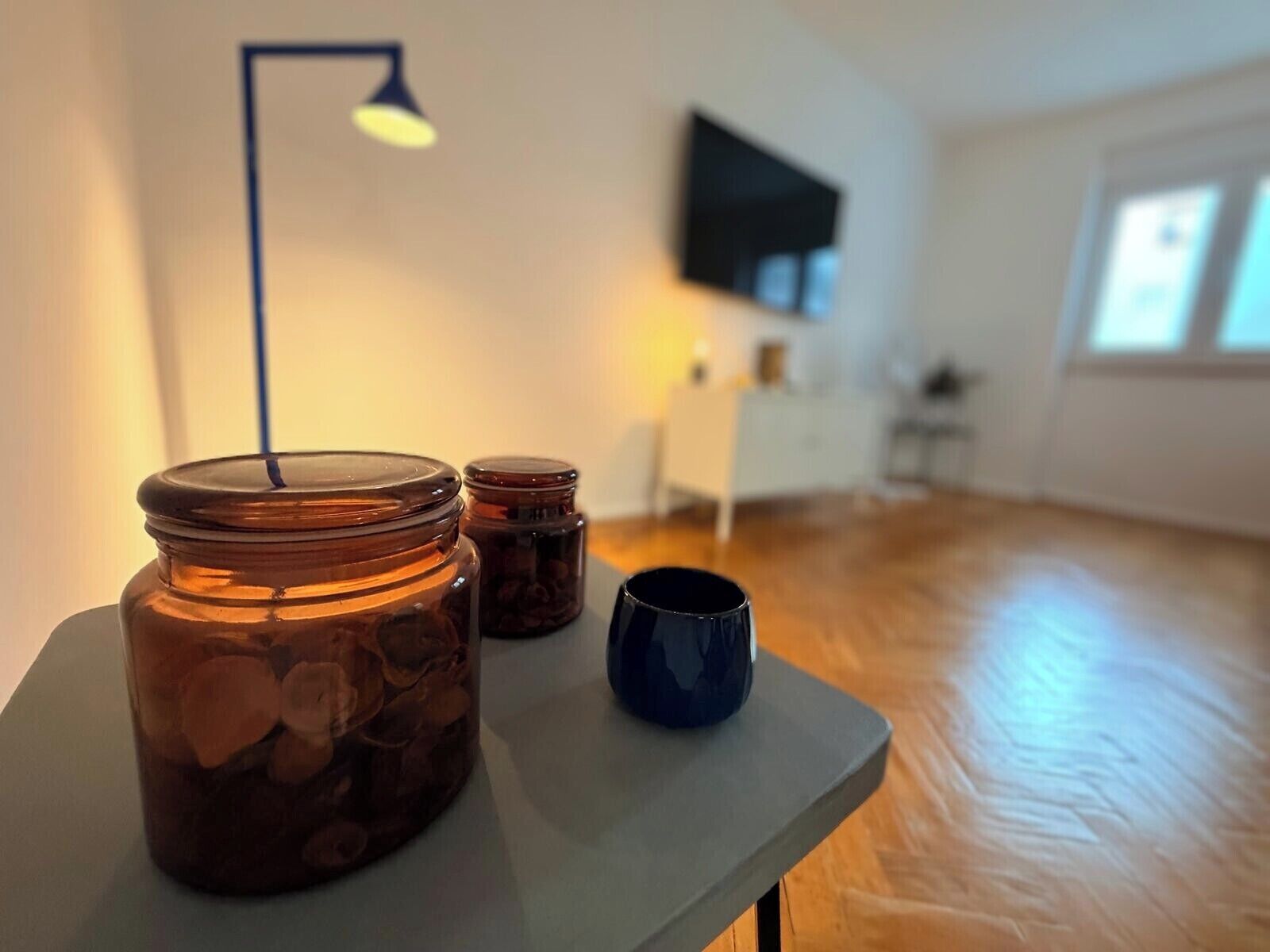 Une pièce avec un plancher en bois, une table sur laquelle se trouvent des bocaux et une tasse, une lampe et un téléviseur fixé au mur.