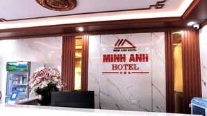 Un comptoir d’accueil avec une enseigne indiquant «Hôtel Minh Anh», un réfrigérateur rempli de boissons et un arrangement floral.