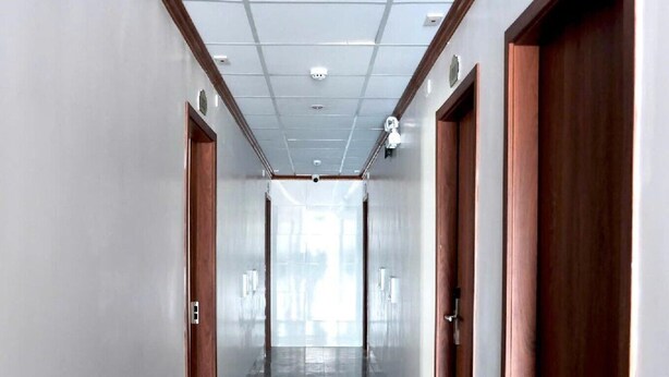 Un long couloir étroit, doté d’un sol carrelé, de portes en bois et d’un plafond pourvu d’un éclairage encastré.