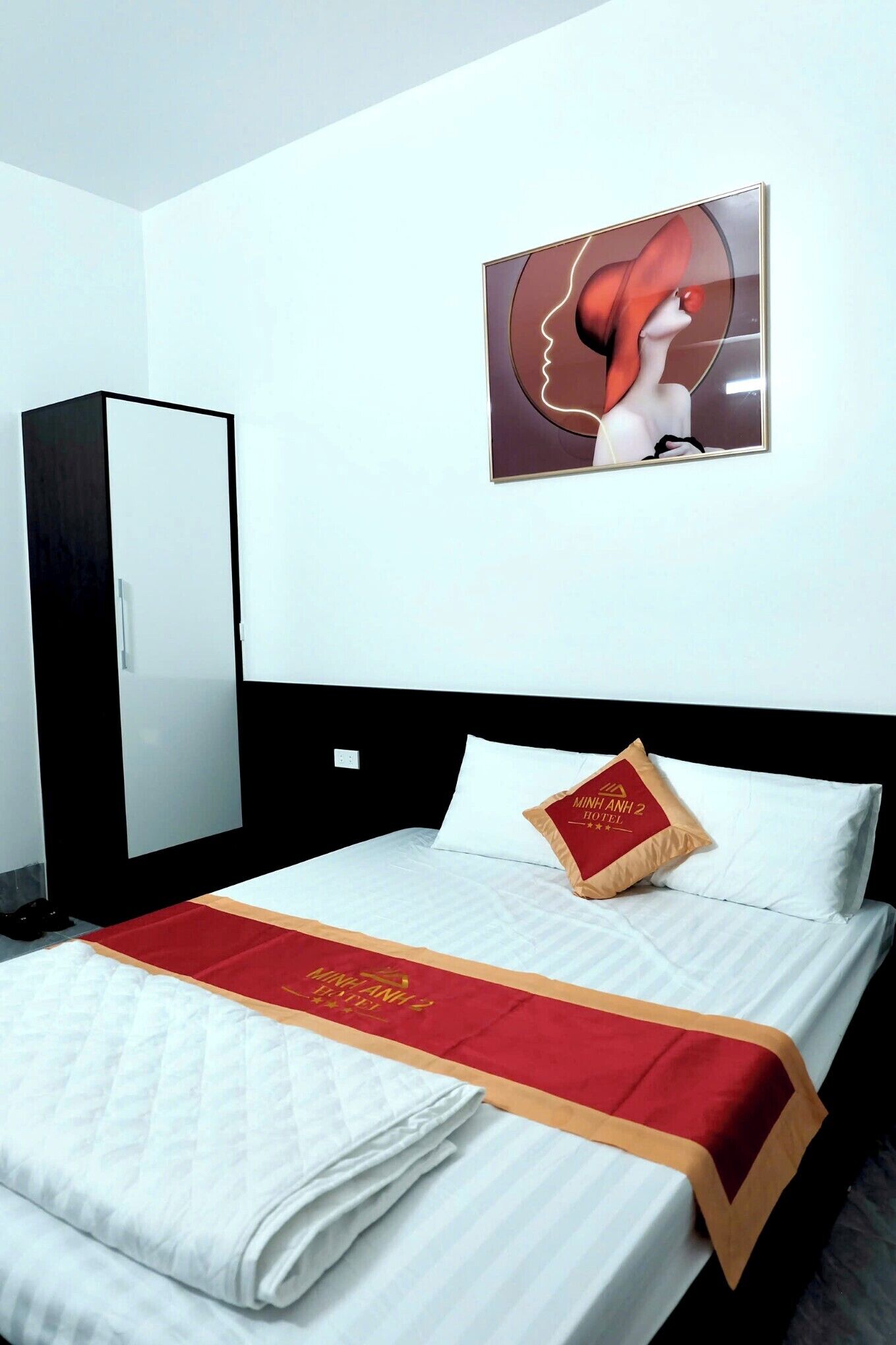 A hotel room with a bed, a red and gold bedspread, a pillow with hotel branding, a wardrobe, and a wall art piece.