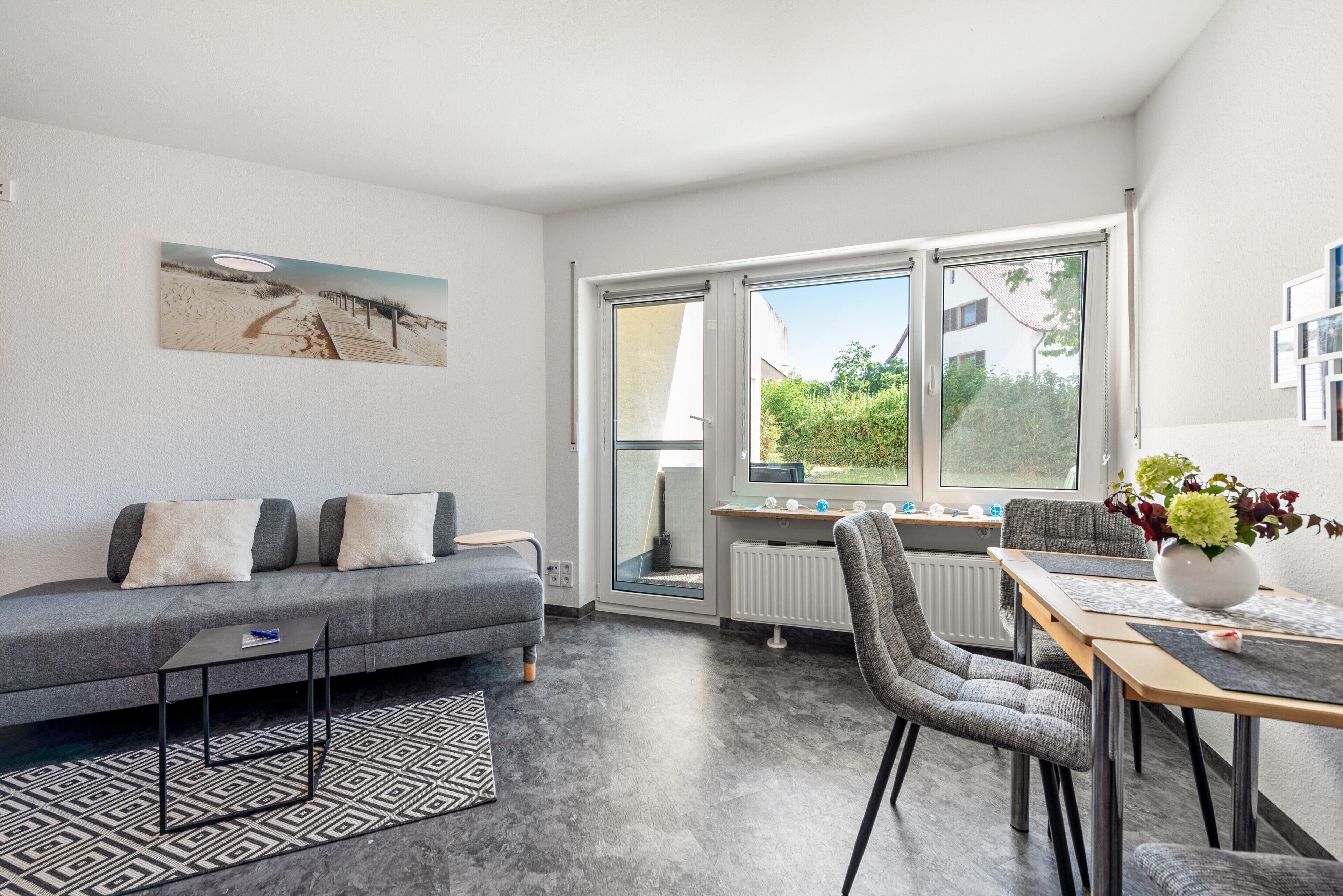 Ein modernes Wohnzimmer mit einem grauen Sofa, einem Esstisch mit Stühlen, einem großen Fenster und einer Heizung.
