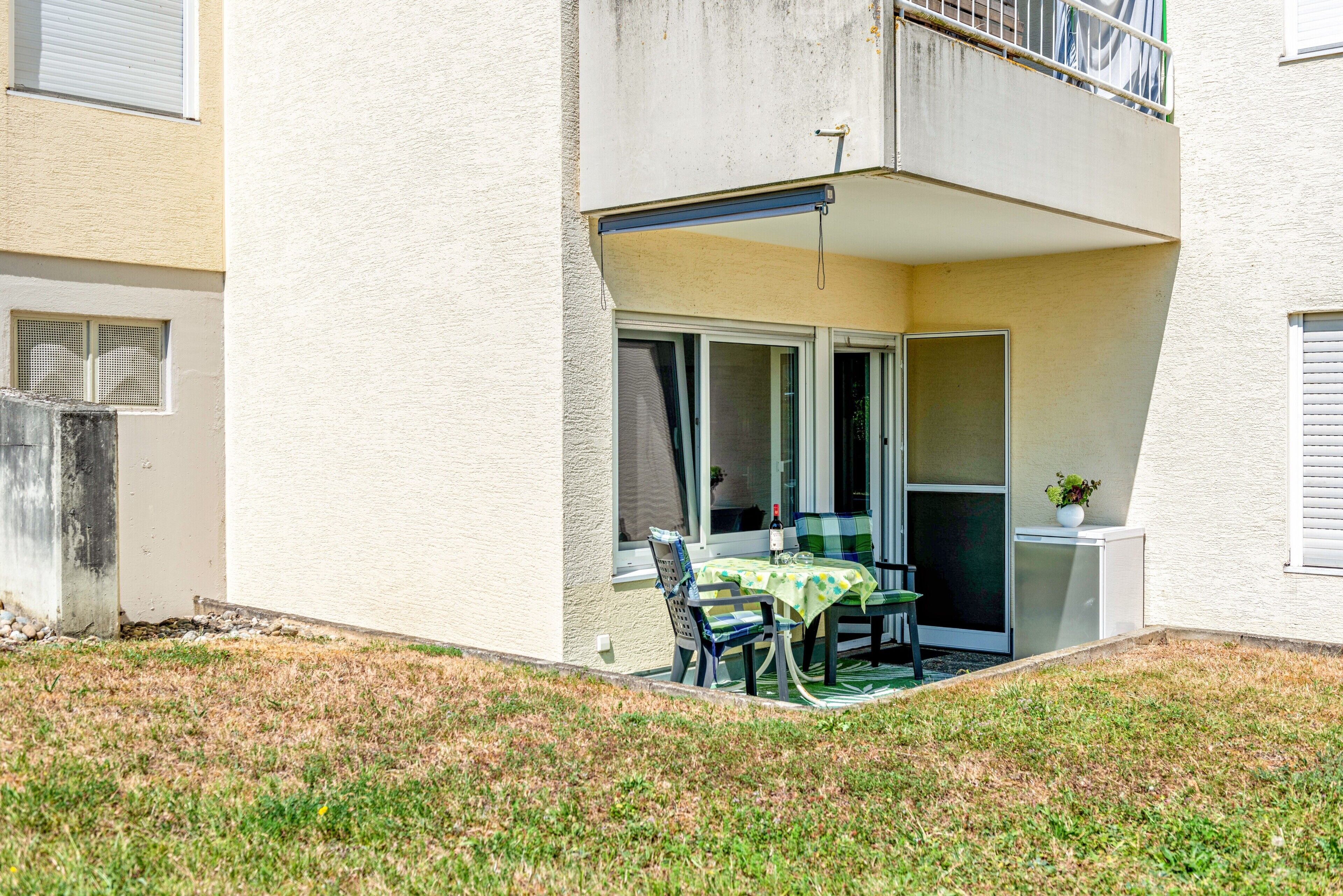 Ein Wohngebäude mit einer überdachten Terrasse, auf der sich ein Tisch, Stühle, ein kleiner Kühlschrank und ein Blumentopf befinden.