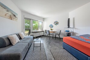 A modern, well-lit room with a grey sofa, a small dining table, and a bed with a striped cover. - Apartment 'Am Hafen' with Lake View, Private Terrace, and Wi-Fi (Konstanz)