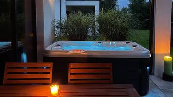 A hot tub with jets, surrounded by wooden chairs and a table with a candle, set against a backdrop of greenery and a building.