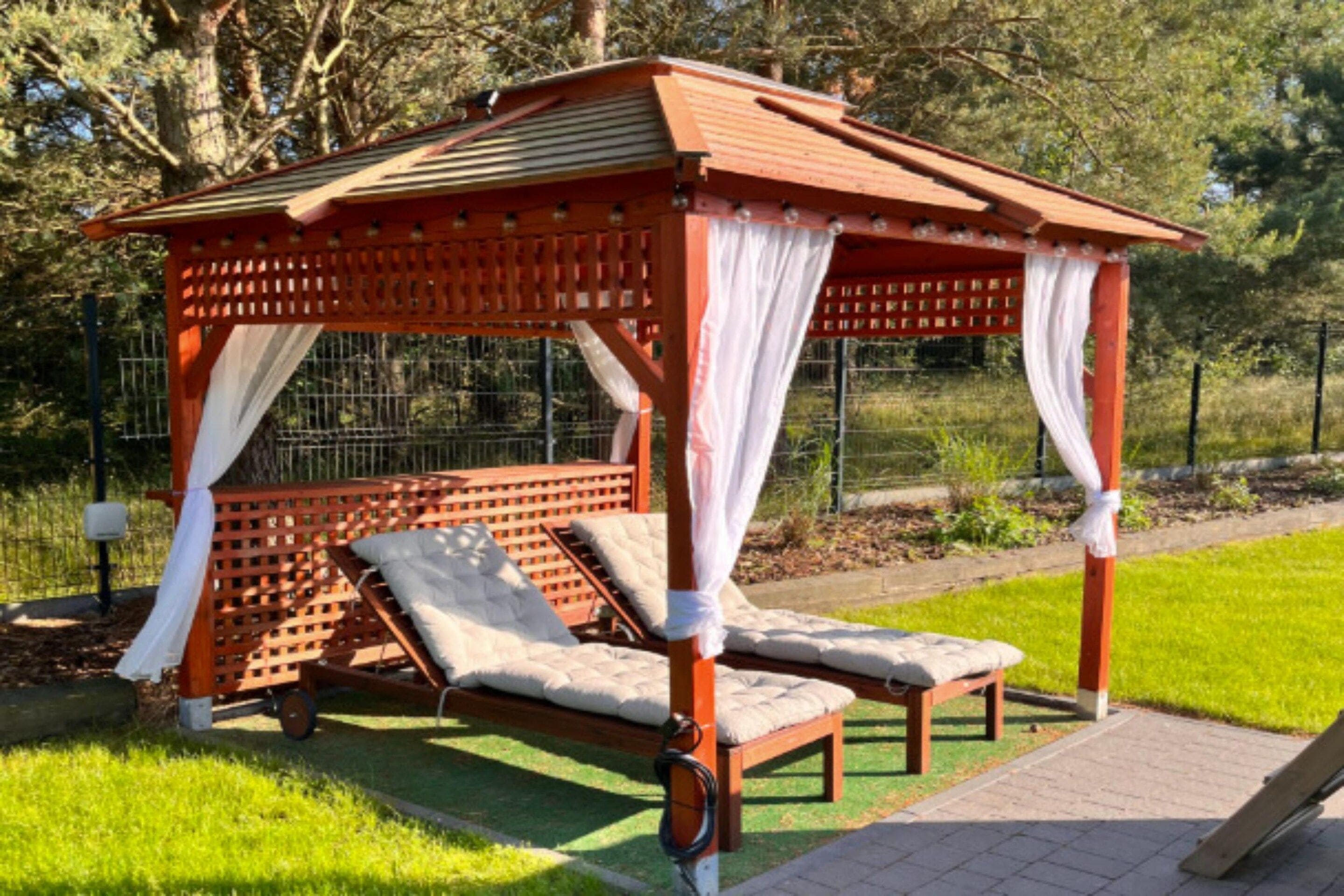 A wooden gazebo with a sloped roof and white curtains, featuring two cushioned benches and a small table.