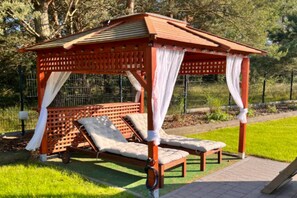 Gazebo in legno con tetto spiovente e tende bianche, dotato di due panche imbottite e un tavolino.